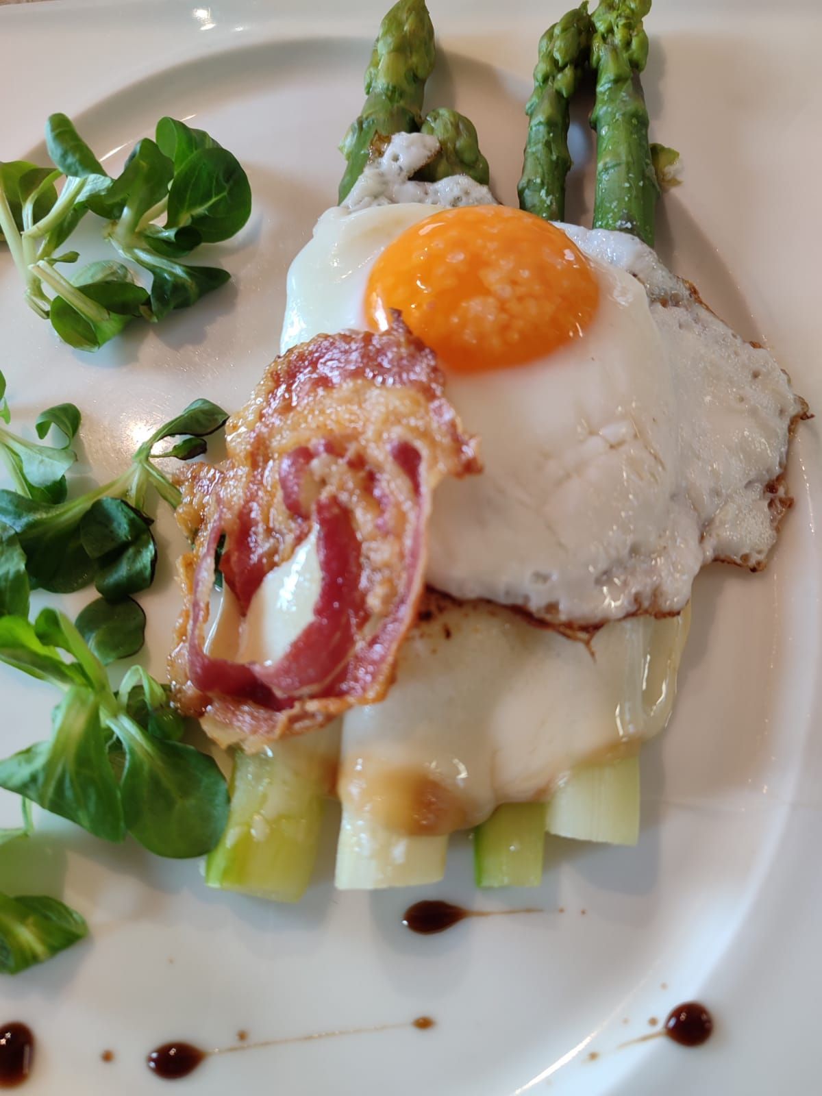 Asparagi, uovo fritto e panino con pancetta su un piatto bianco, guarnito con verdure e un filo di aceto balsamico.