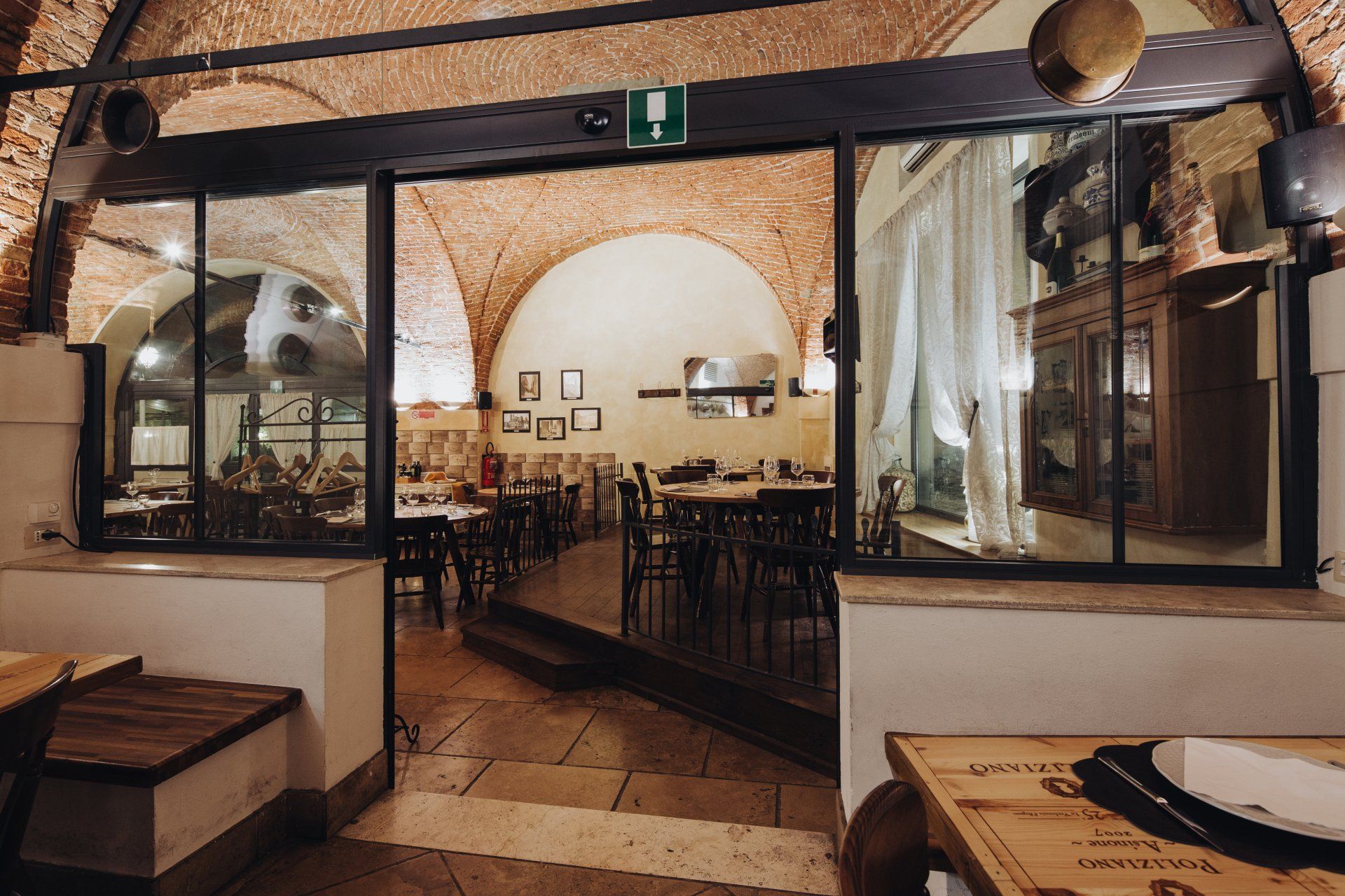 Interno del ristorante con soffitti a volta in mattoni, tavoli, sedie e una porta con un cartello verde di uscita.