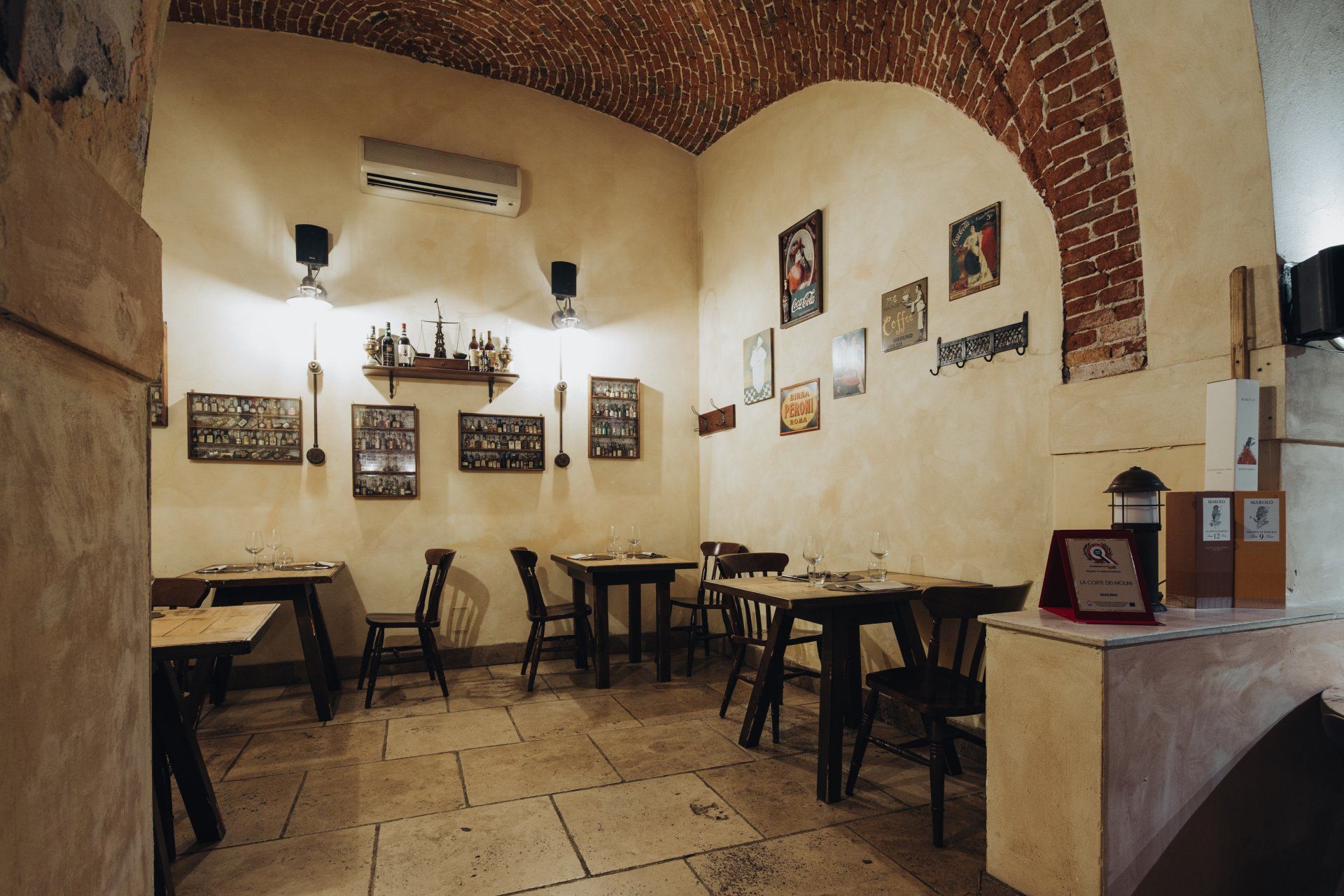 Interno di un piccolo ristorante con soffitto ad arco in mattoni, pavimento in pietra, tavoli, sedie e quadri incorniciati alle pareti.