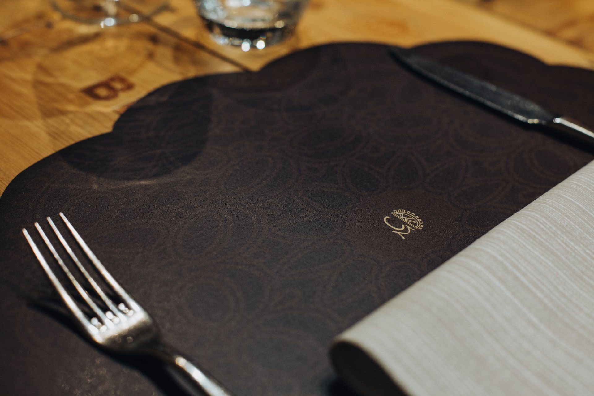 Primo piano di una tovaglietta, con forchetta, coltello e tovagliolo piegato. Marrone scuro, bordo smerlato.