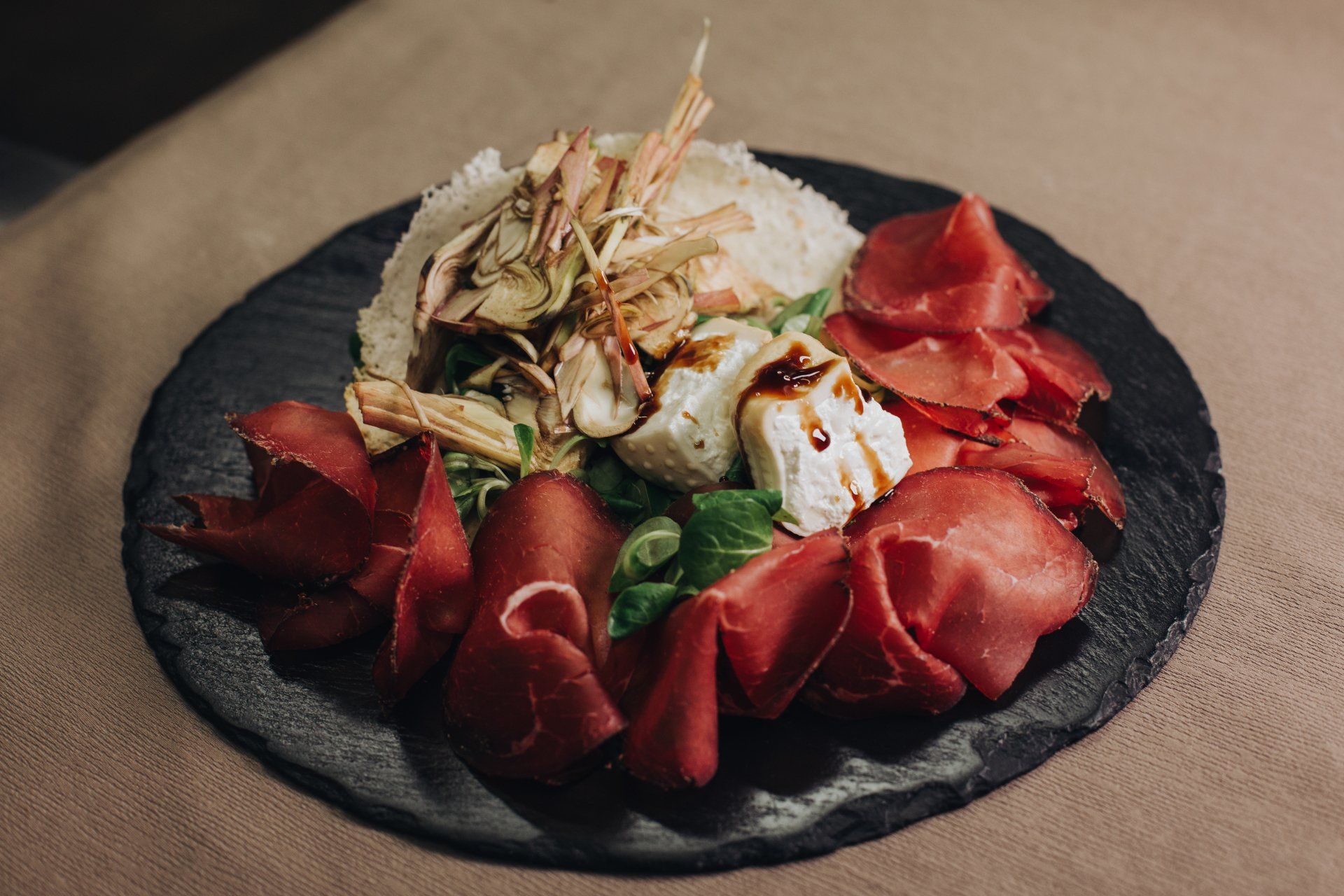 Piatto di antipasti: salumi, formaggi, cuori di carciofo e verdure, disposti su un piatto di pietra scura.