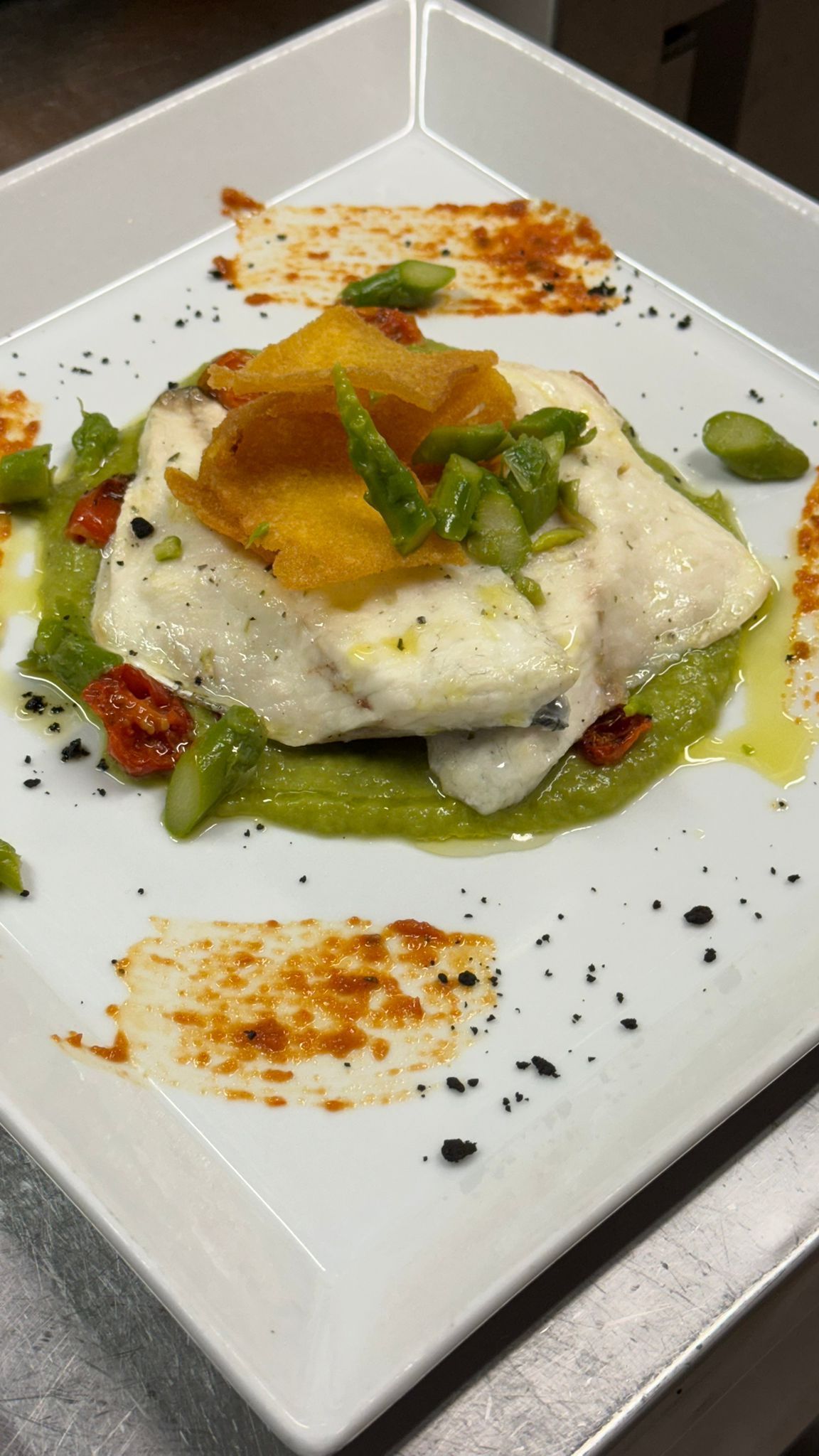 Filetto di pesce bianco con asparagi, salsa e guarnizione arancione su un piatto bianco.