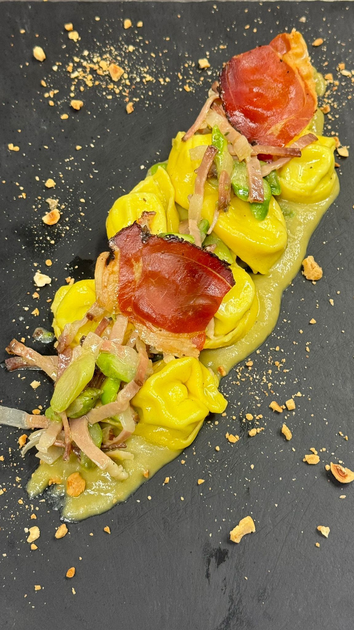 Tortellini gialli con prosciutto, verdure e salsa su un piatto nero.