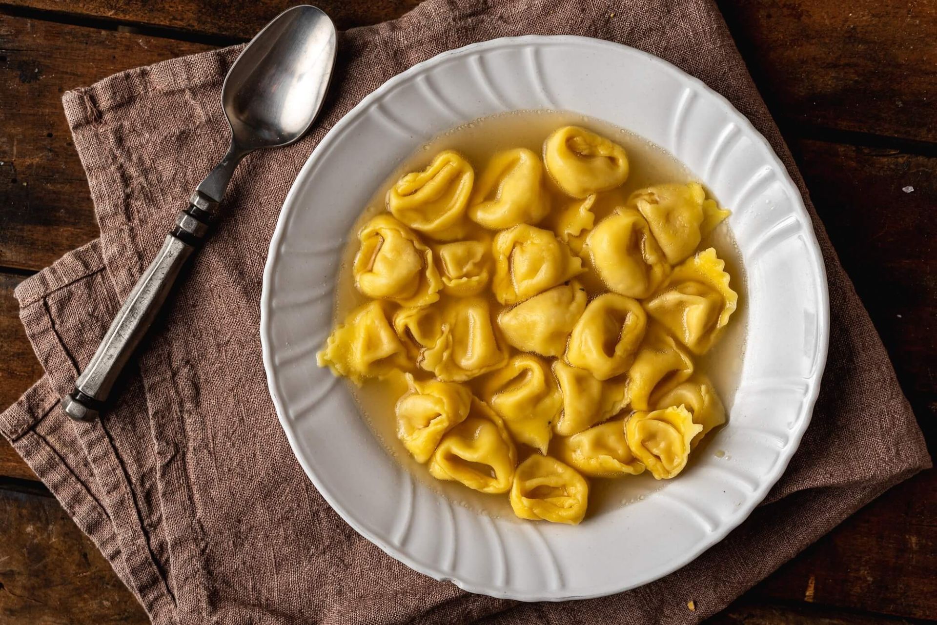 Zuppa di tortellini in una ciotola bianca, su un tovagliolo marrone, con un cucchiaio.
