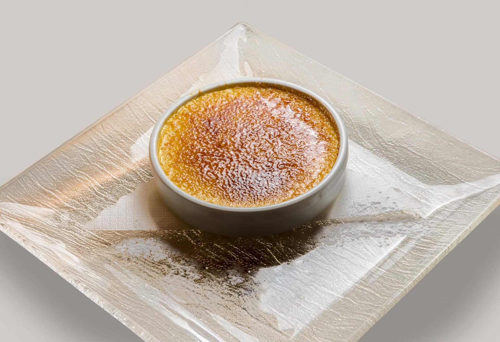 Crème brûlée in una piccola cocotte di metallo su un piatto di vetro quadrato, spolverata con zucchero a velo.