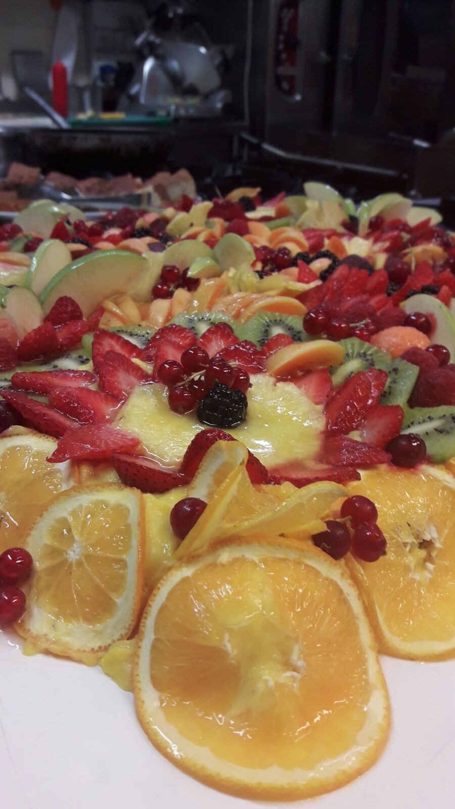 Primo piano di una crostata di frutta colorata con arance, fragole, kiwi e frutti di bosco disposti su una superficie bianca.