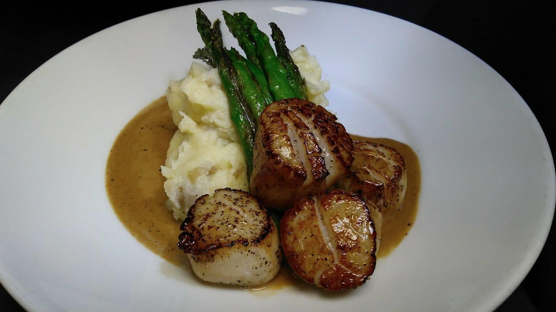 Seared scallops with mashed potatoes, asparagus, and gravy on a white plate