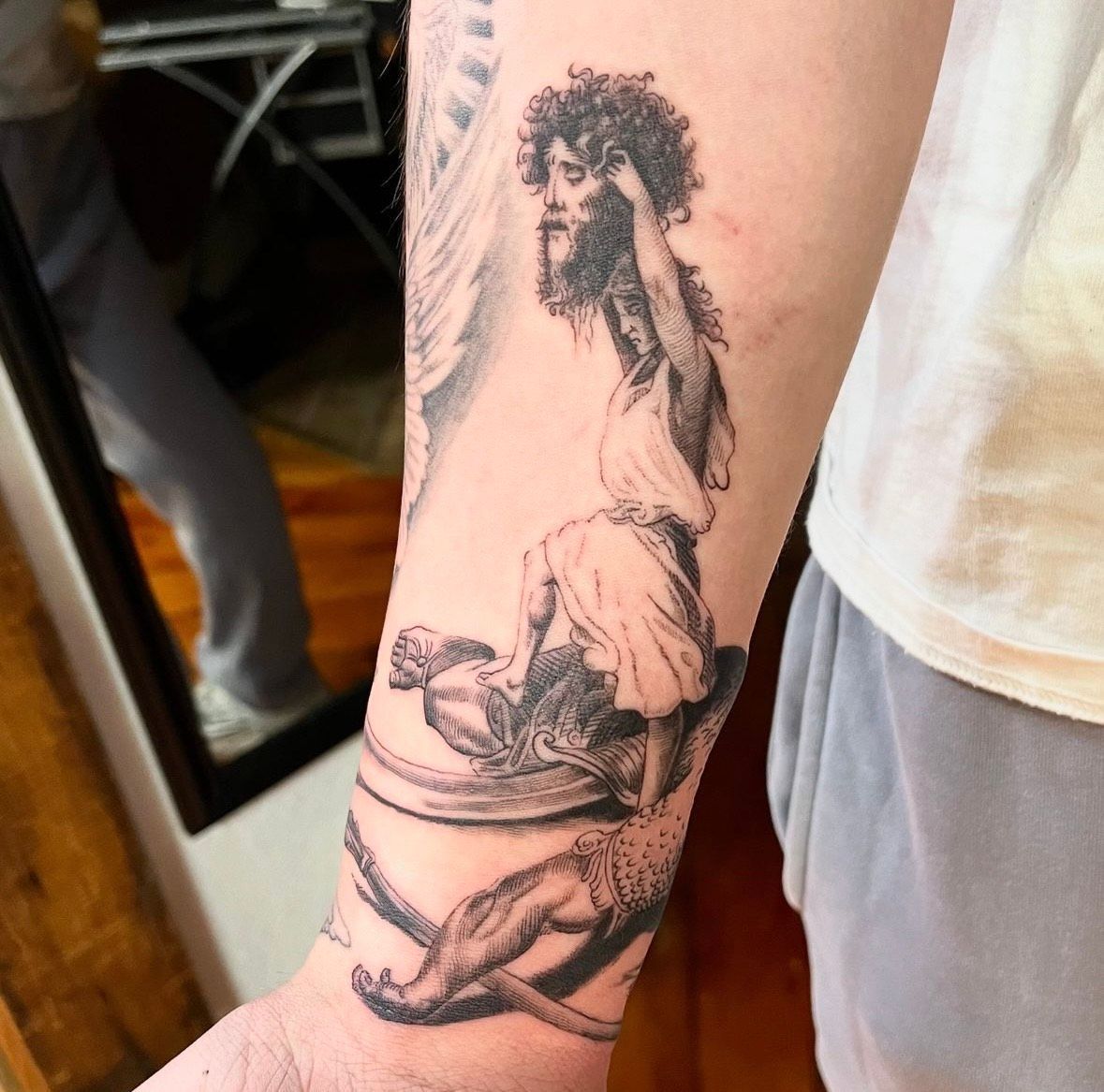Black and white tattoo on arm depicting a figure atop another, possibly in a classical style.