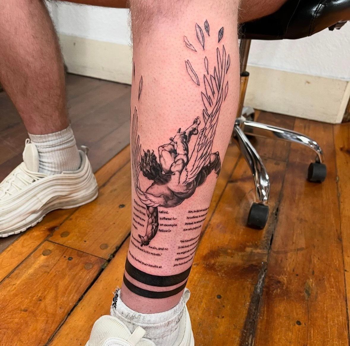 Tattoo on a leg featuring a person with wings, text below, and black bands around the ankle, against a wooden floor.