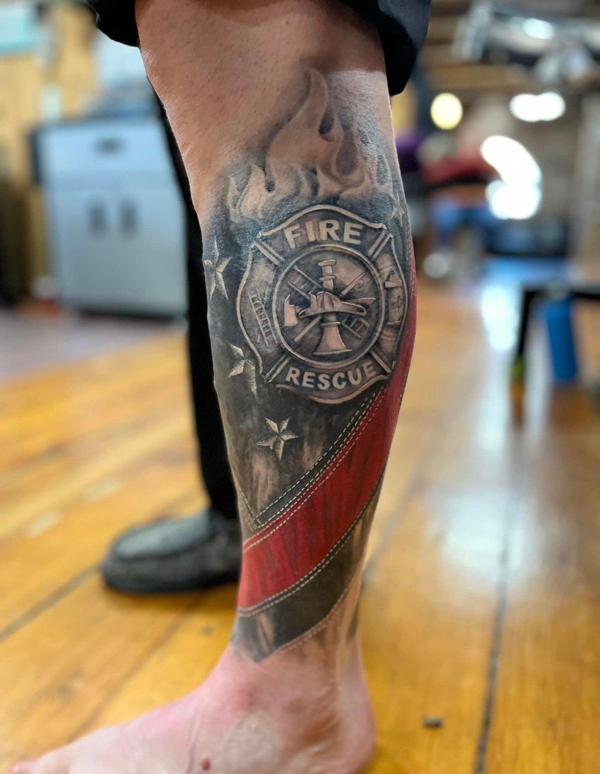 Firefighter's calf tattoo: fire department emblem, red line on flag, flames, gray and black shading.