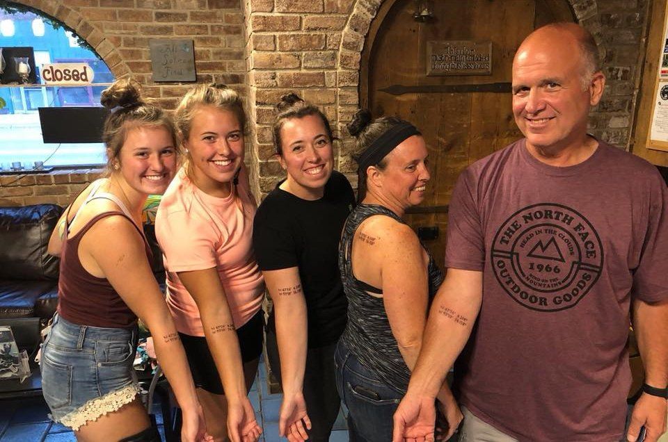 Five people with matching arm tattoos pose together; brick wall background.