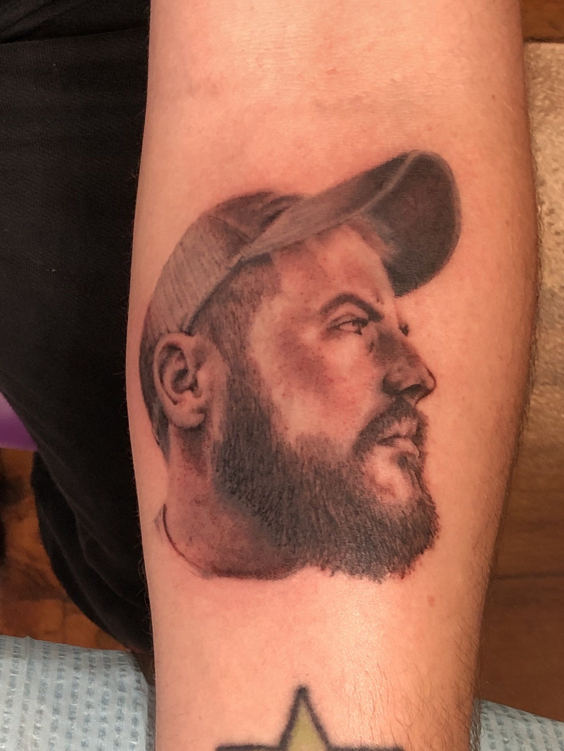 Tattoo of a bearded person in a baseball cap, in profile, on a person's arm. Black and gray shading.