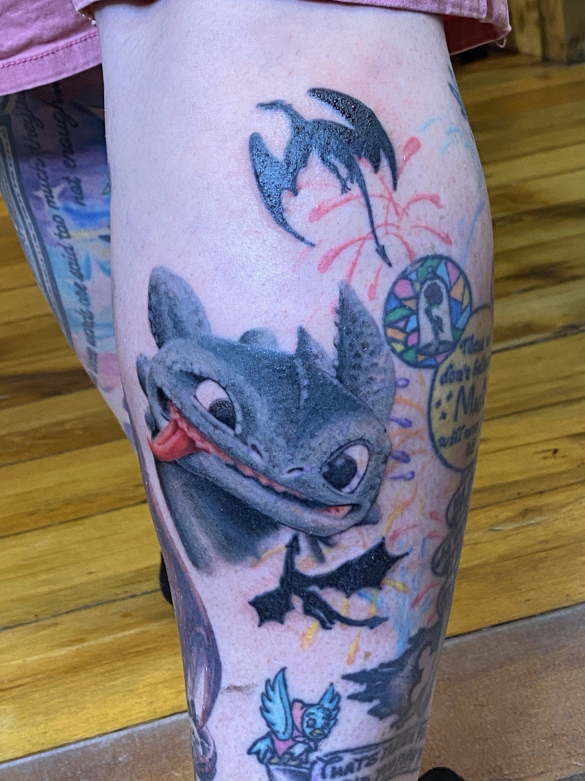 Tattoo of Toothless the dragon sticking out his tongue on a calf. Other small animated images and fireworks.