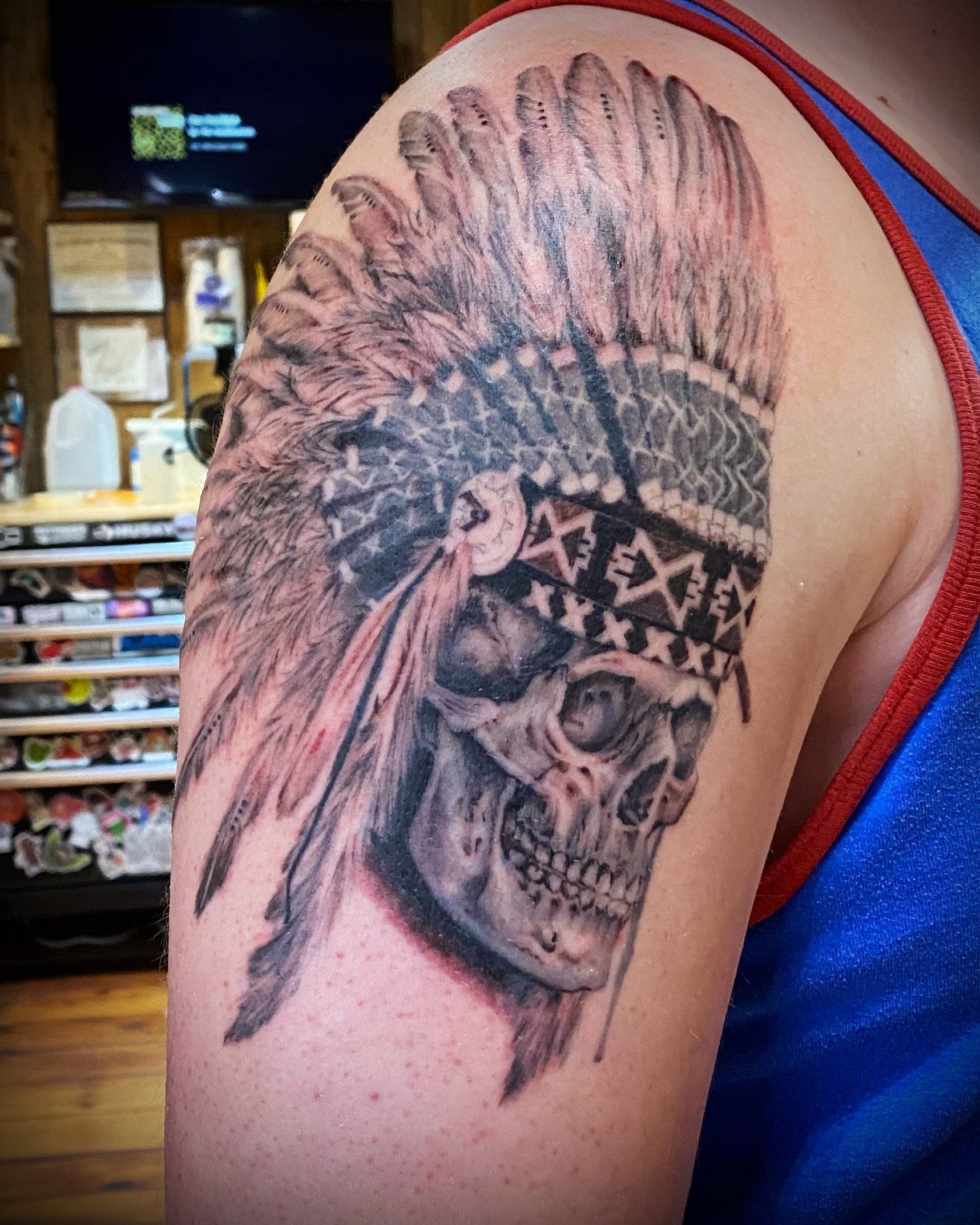 Tattoo of a skull wearing a Native American headdress on a person's shoulder. Black and gray ink.