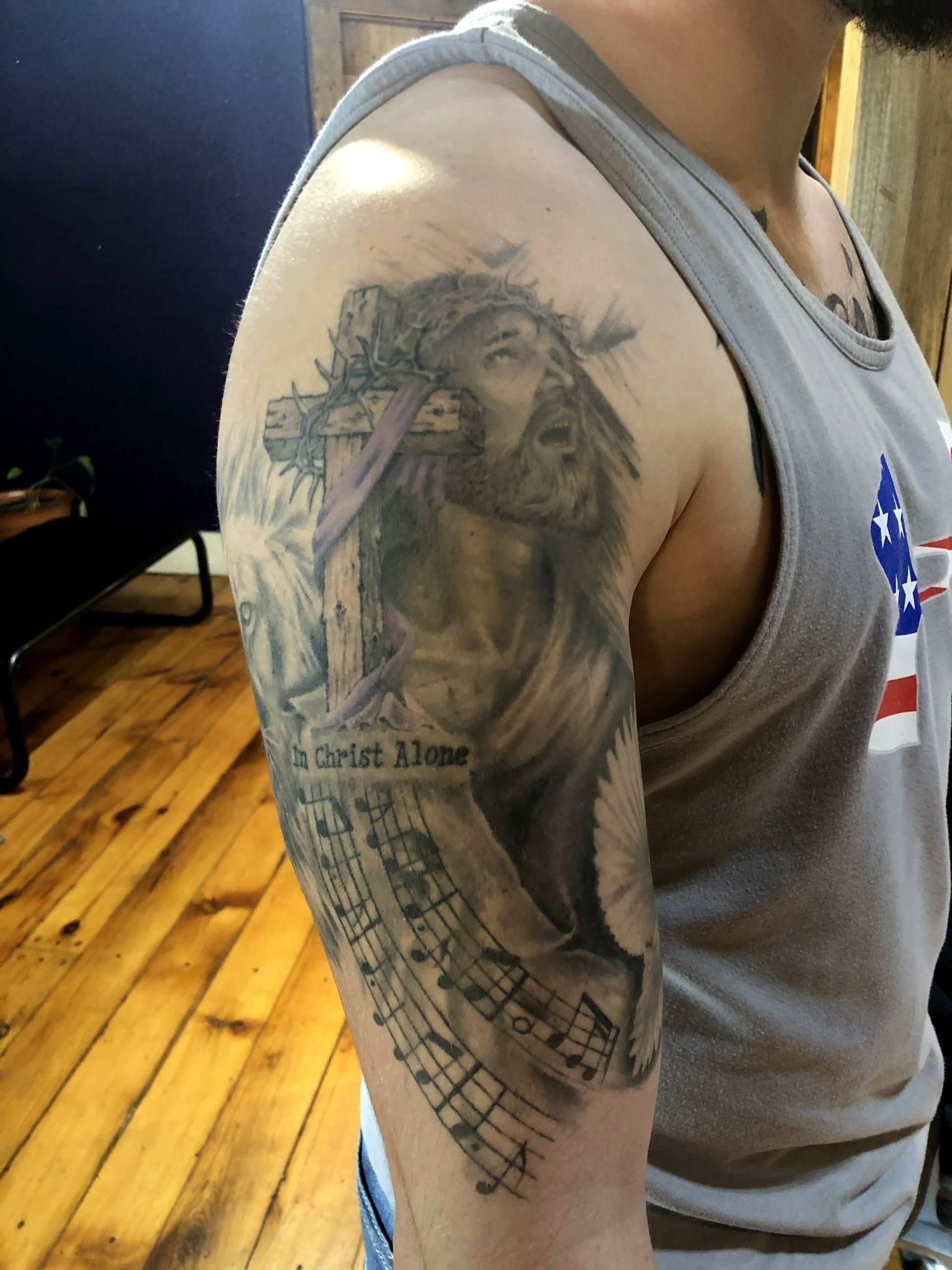 Tattoo of Jesus on a person's upper arm, with text and musical notes. Person wears a gray tank top.