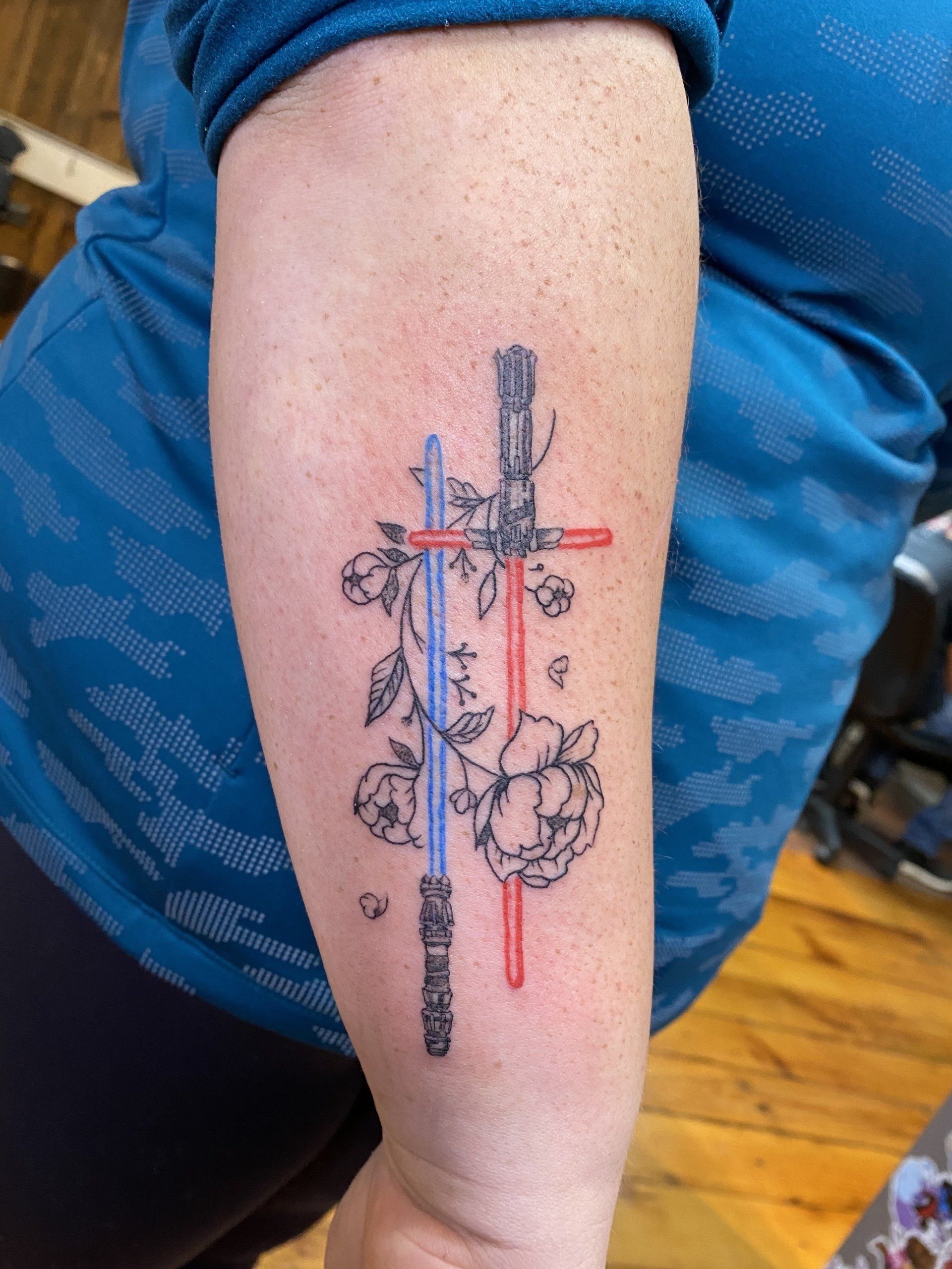 Tattoo of lightsabers crossed, roses, on a person's forearm; lightsabers are red and blue.