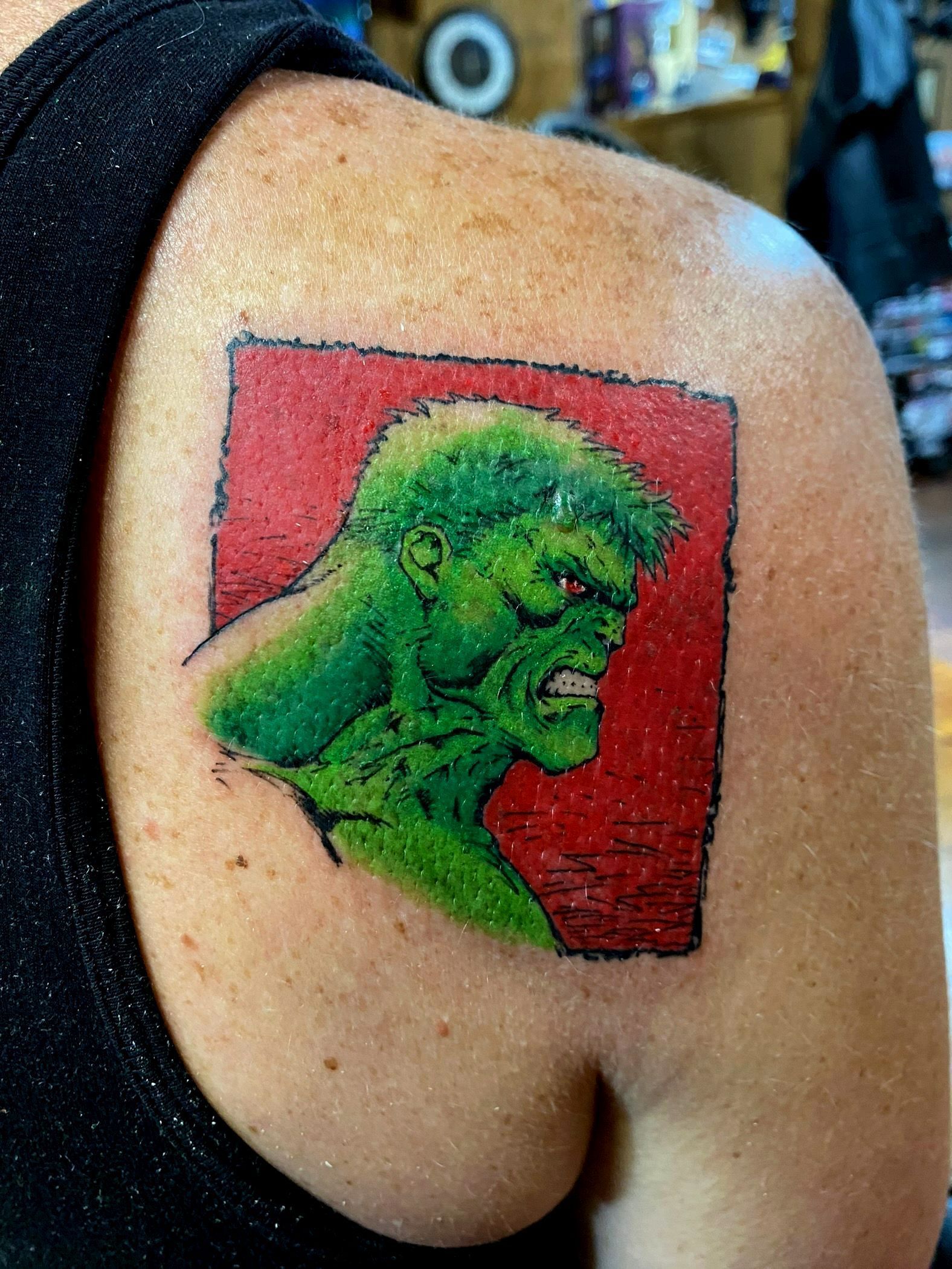 Hulk tattoo on shoulder, green head in red square, angry expression.