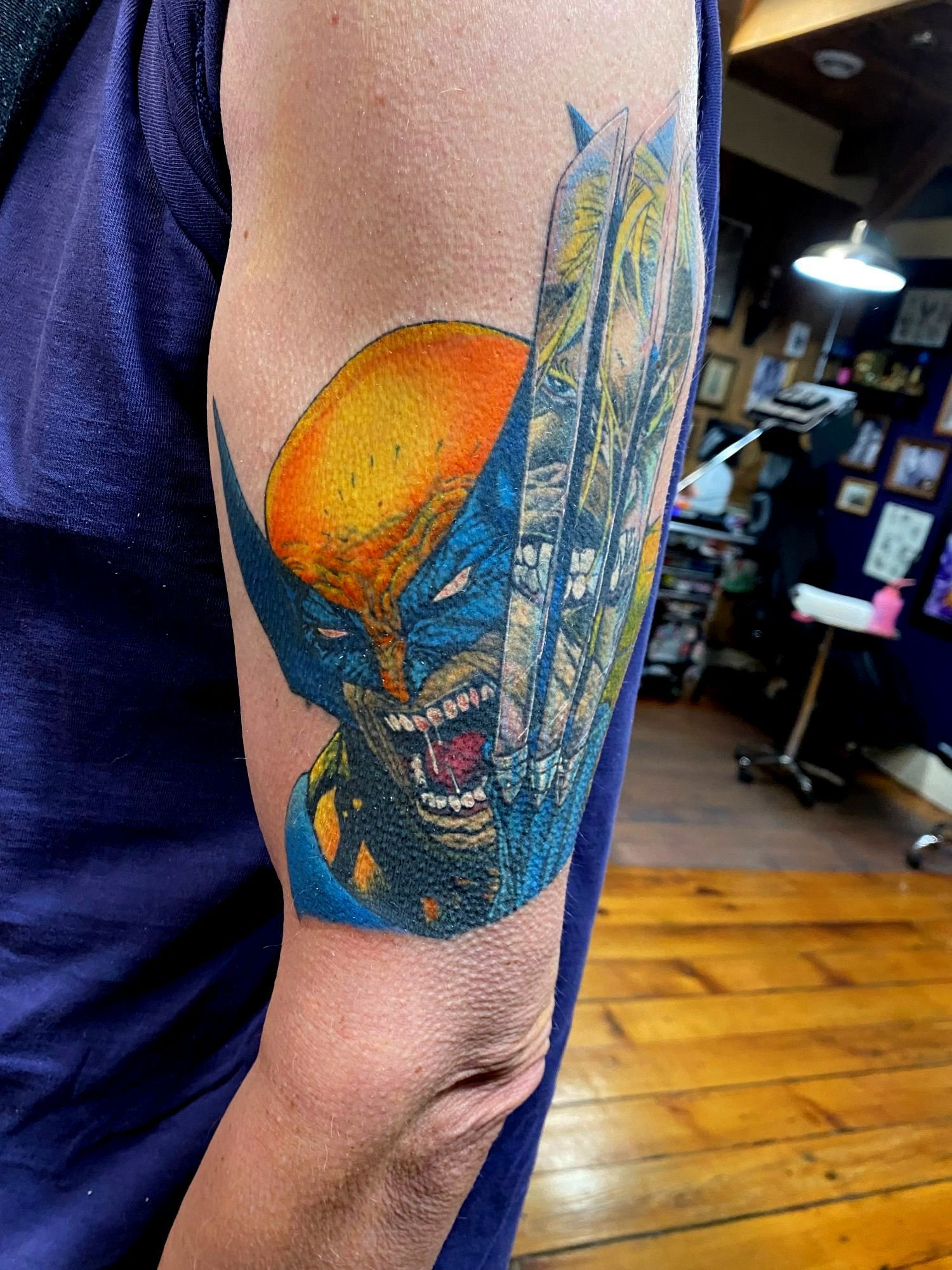 Tattoo of Wolverine's face and claws on a person's upper arm, with blue, yellow, and red colors.