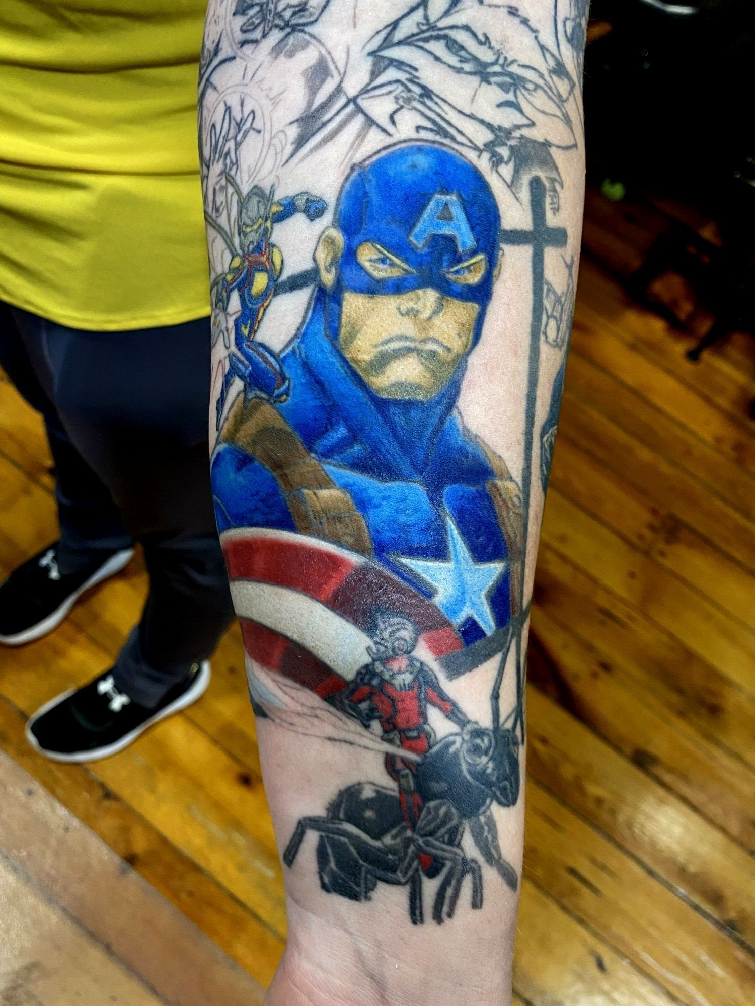 Tattoo of Captain America with Ant-Man. Blue and red, stern face, arm tattoo, indoor setting.