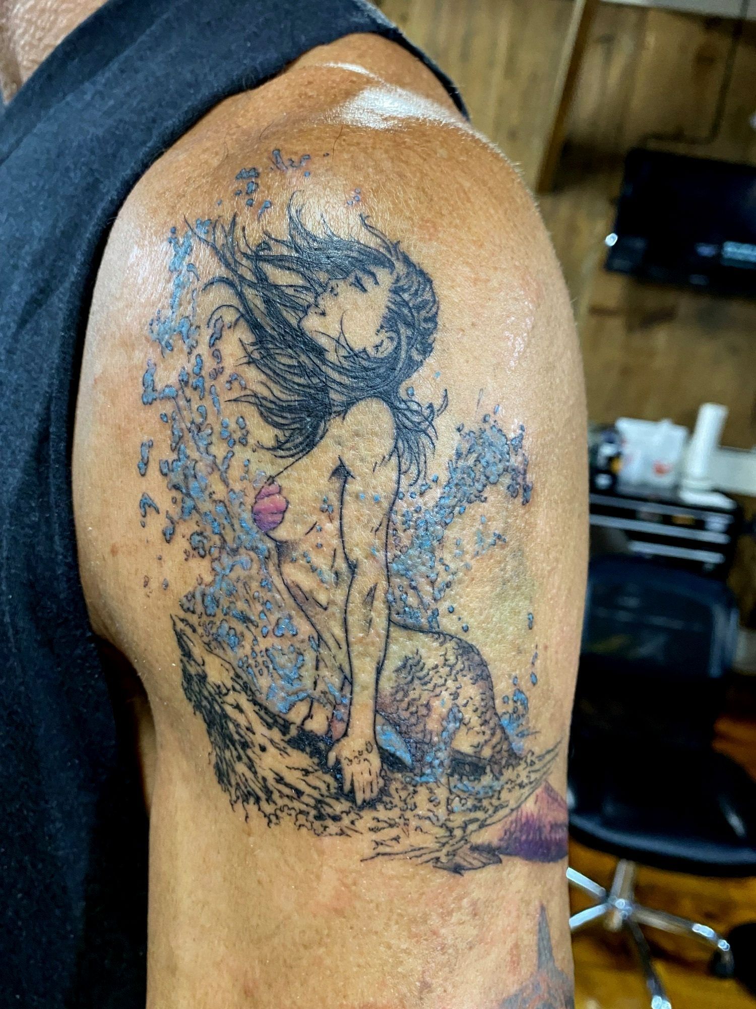 Tattoo of a nude woman with long hair, surrounded by blue and gray splatters, on a person's shoulder.