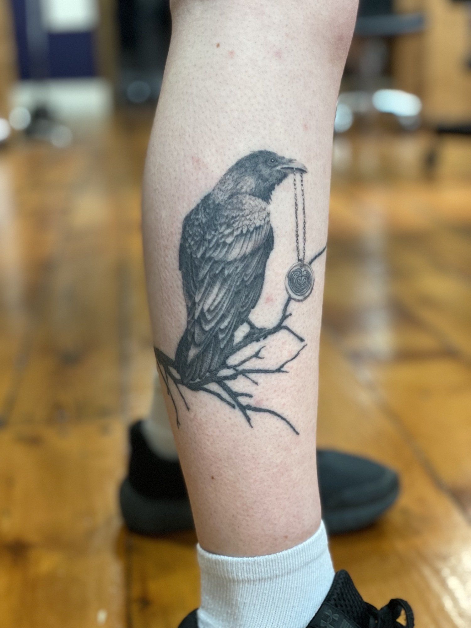 Black crow tattoo holding a pendant on a branch.