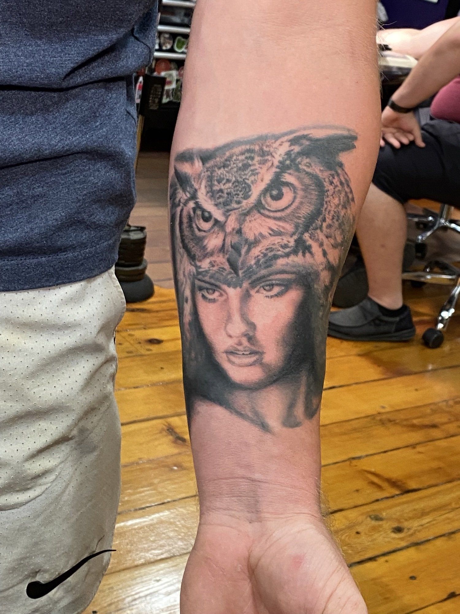 Forearm tattoo: Owl head over a woman's face, black ink.