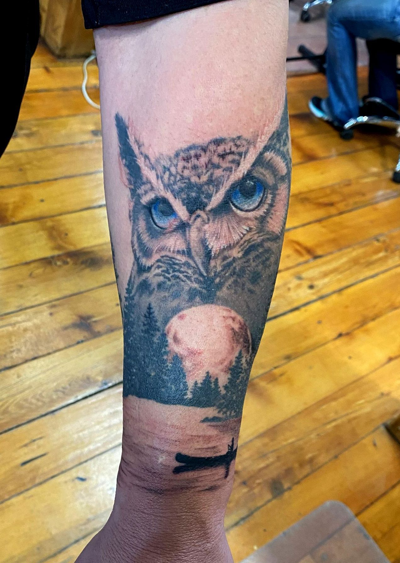 Tattoo of an owl with blue eyes and a forest scene. Done on a person's forearm.
