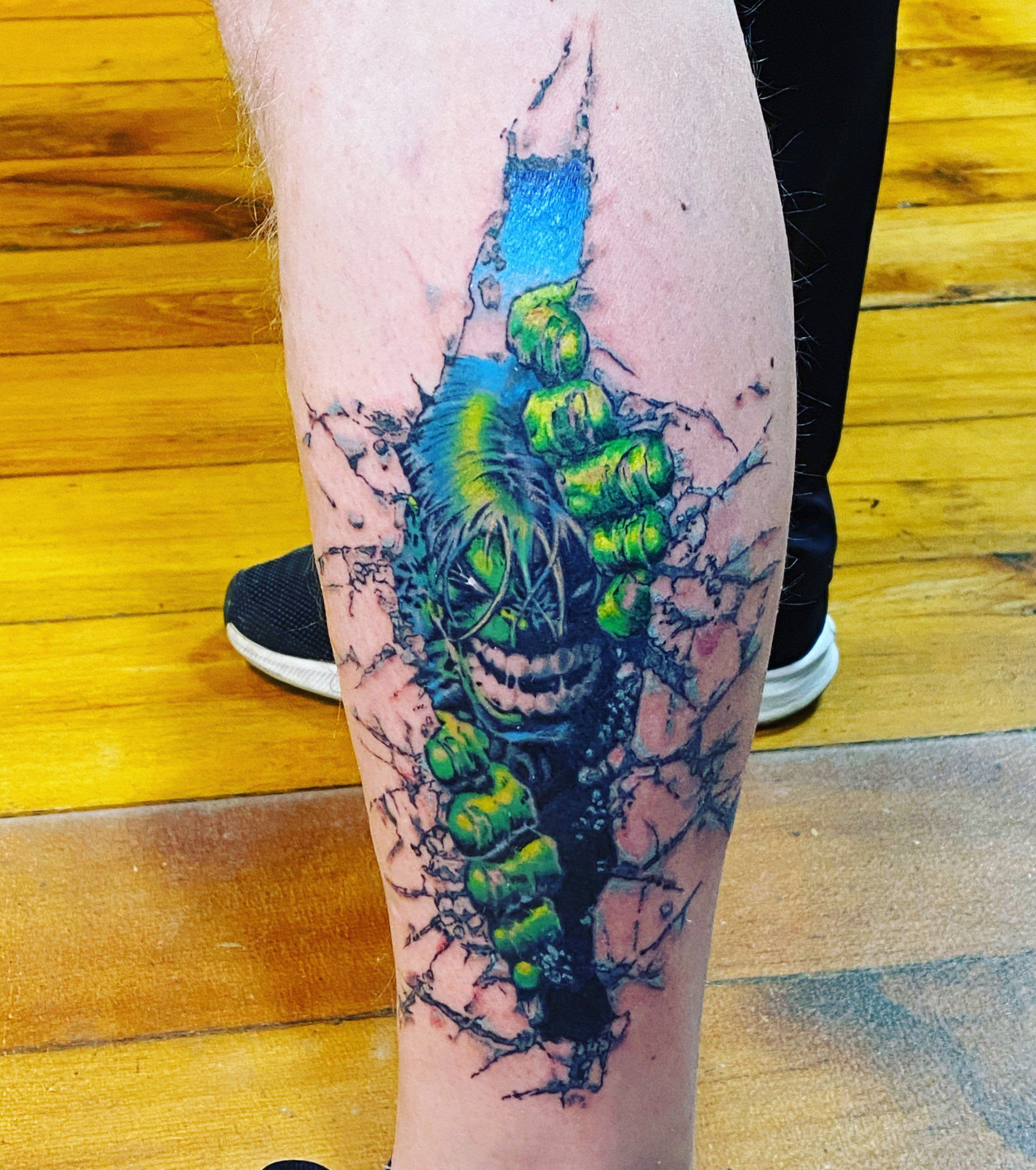 Tattoo of the Hulk bursting through a torn wall, green and blue colors, on a leg.