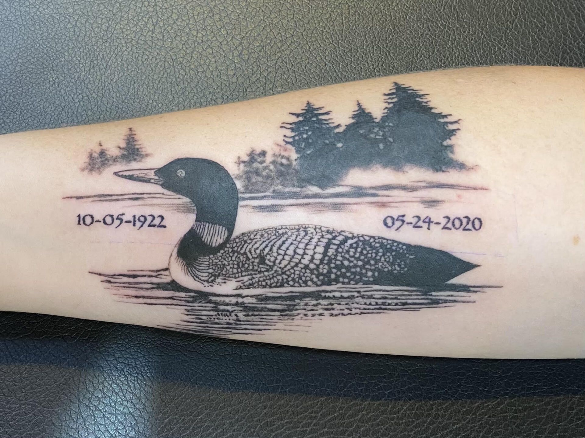 Tattoo of a loon floating on water, with dates 10-05-1922 and 05-24-2020. Trees and a lake are in the background.