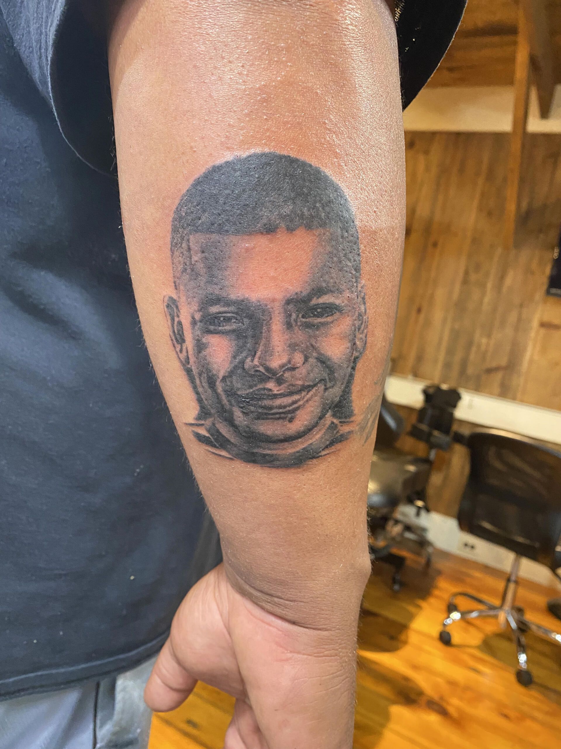 Black and white tattoo of a person’s face on a forearm. Smiling expression, short hair.