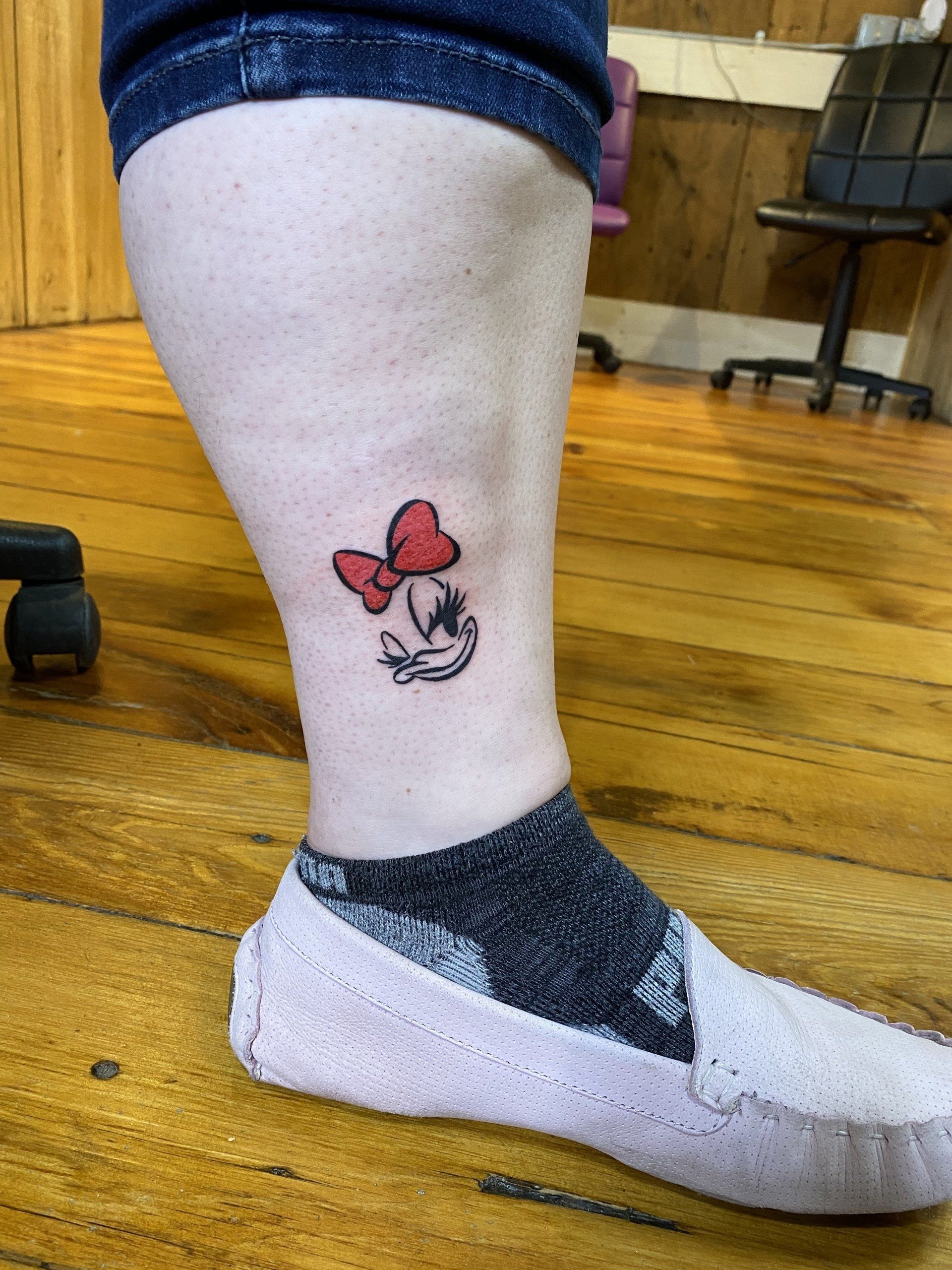 A Minnie Mouse tattoo on a person's ankle with a red bow. The person wears a sock and a light pink shoe.