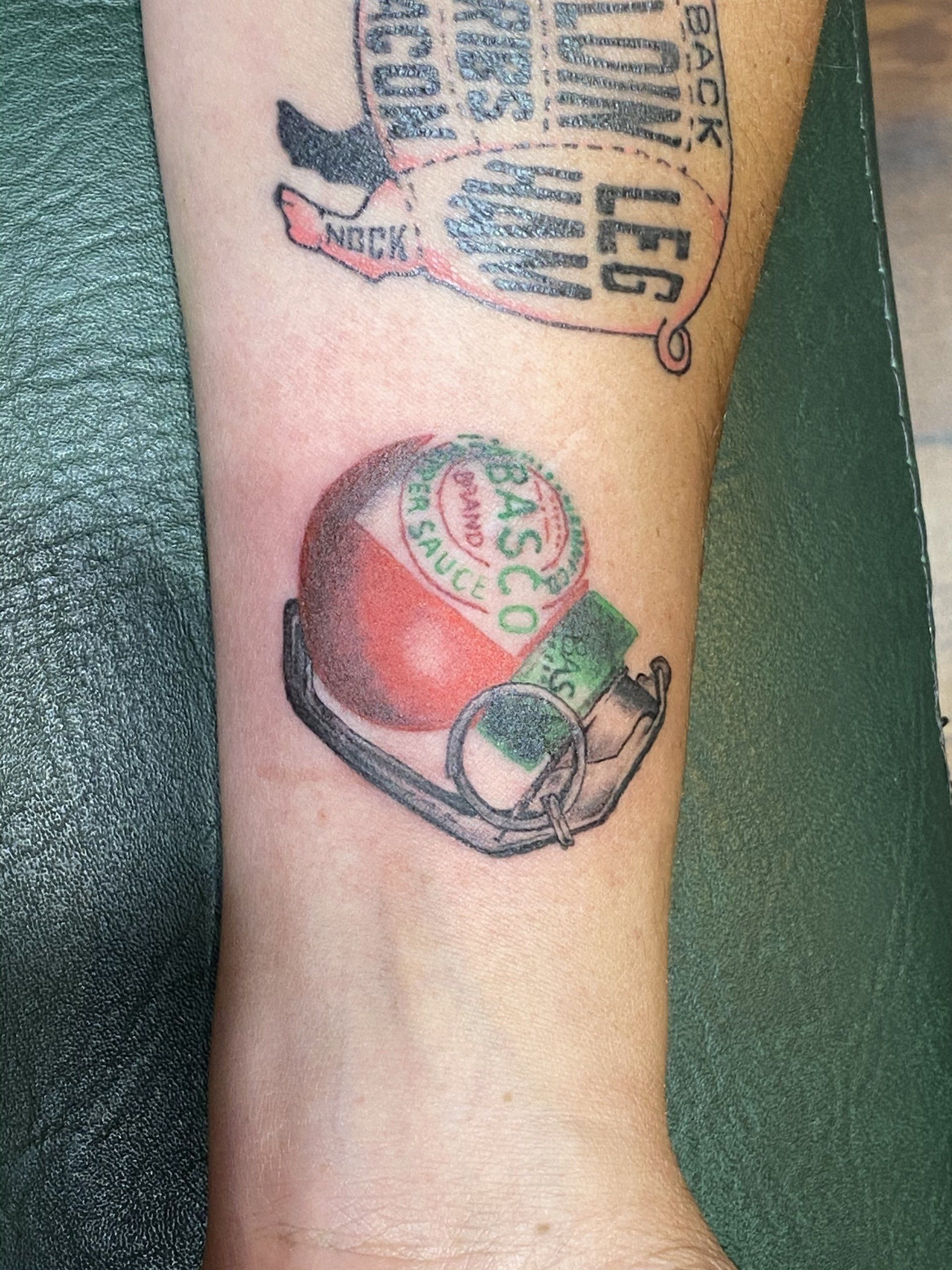 Tattoo of a Tabasco sauce bottle on a person's arm. Above is a meat cuts diagram tattoo.