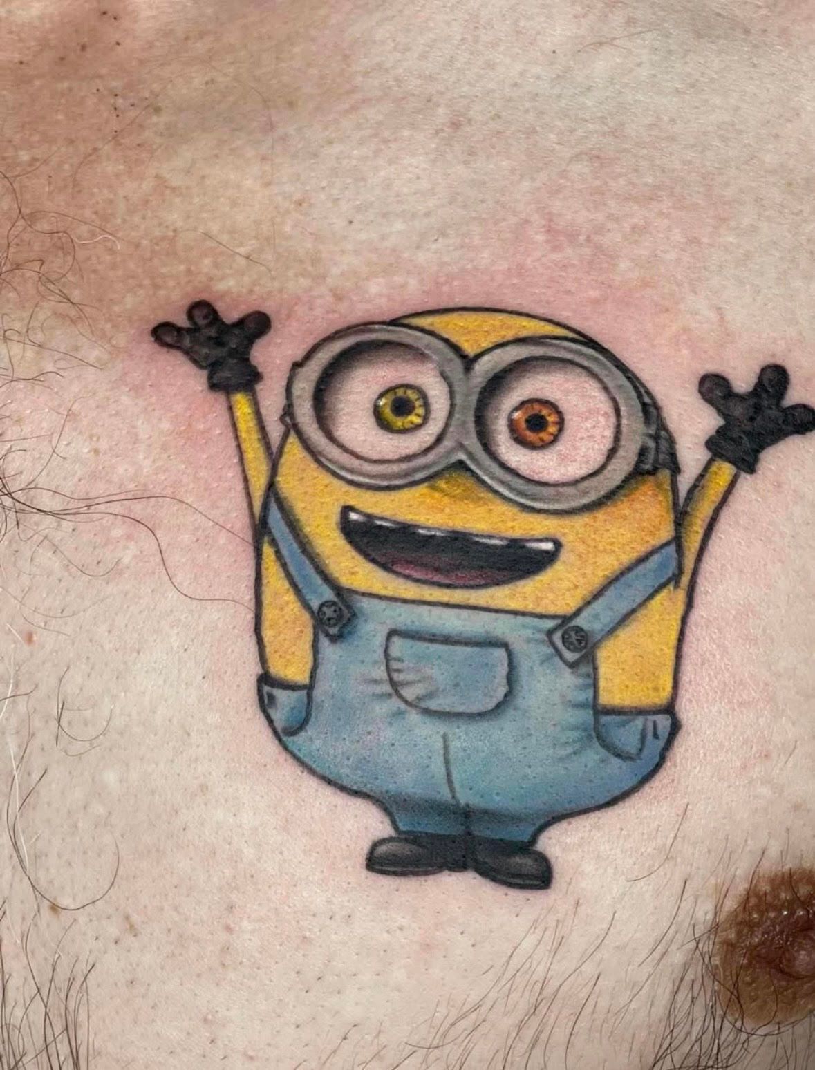 Minion tattoo on chest, yellow and blue, arms raised, smiling.