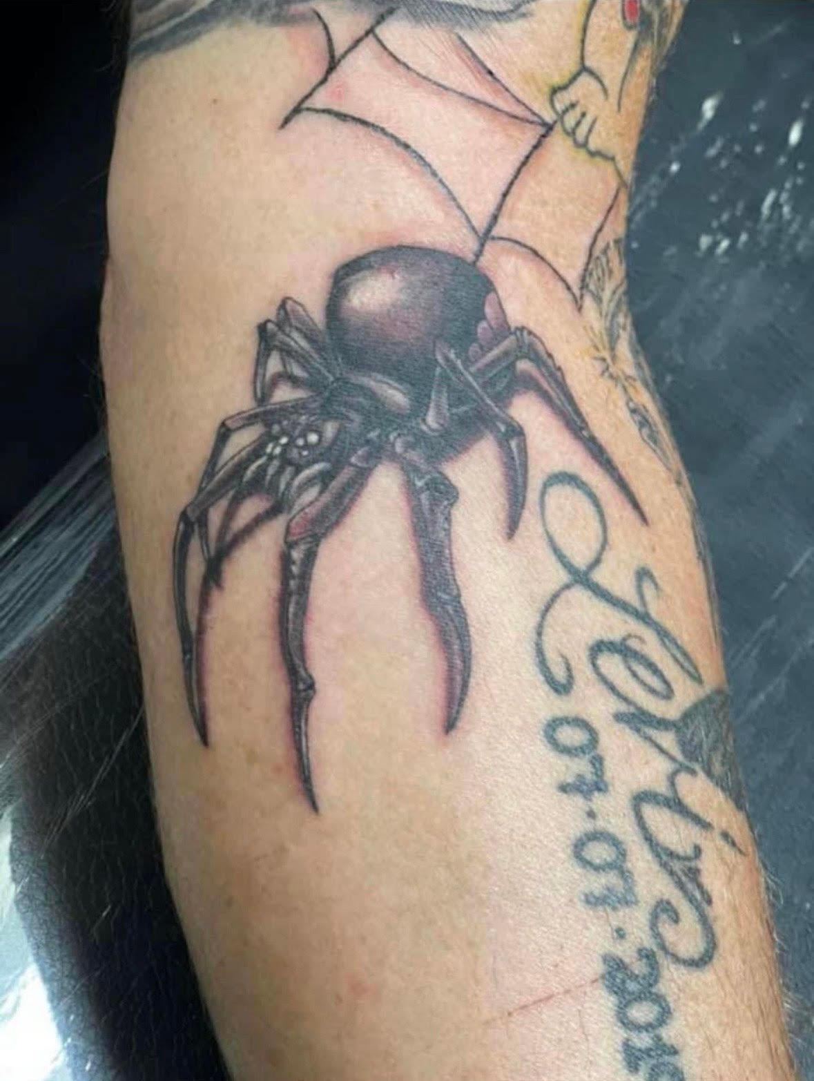 Tattoo of a black spider on an arm with a partial web and script.
