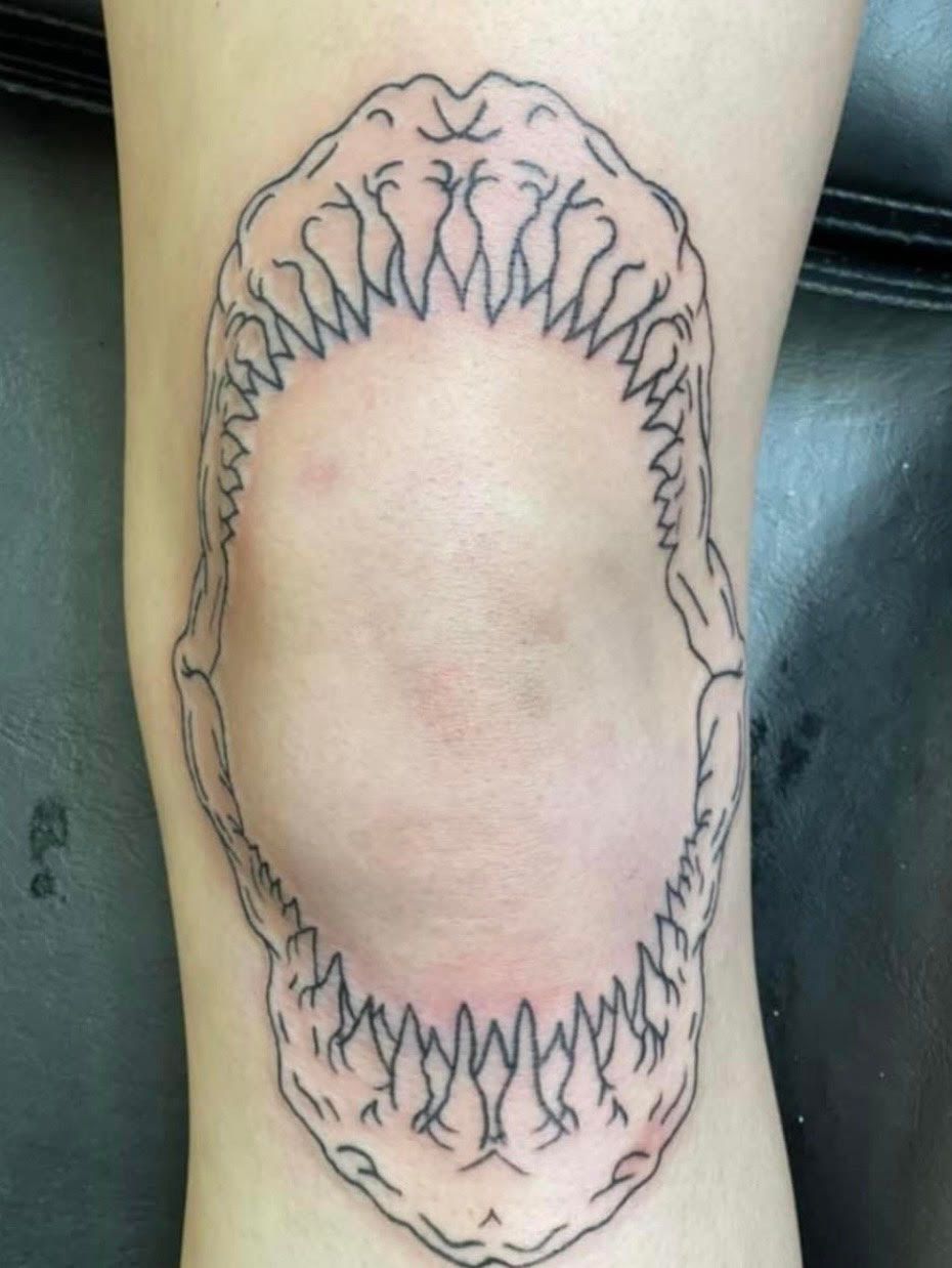 Shark jaws tattoo on a knee, outlined in black ink. Teeth point inward, framing the skin.