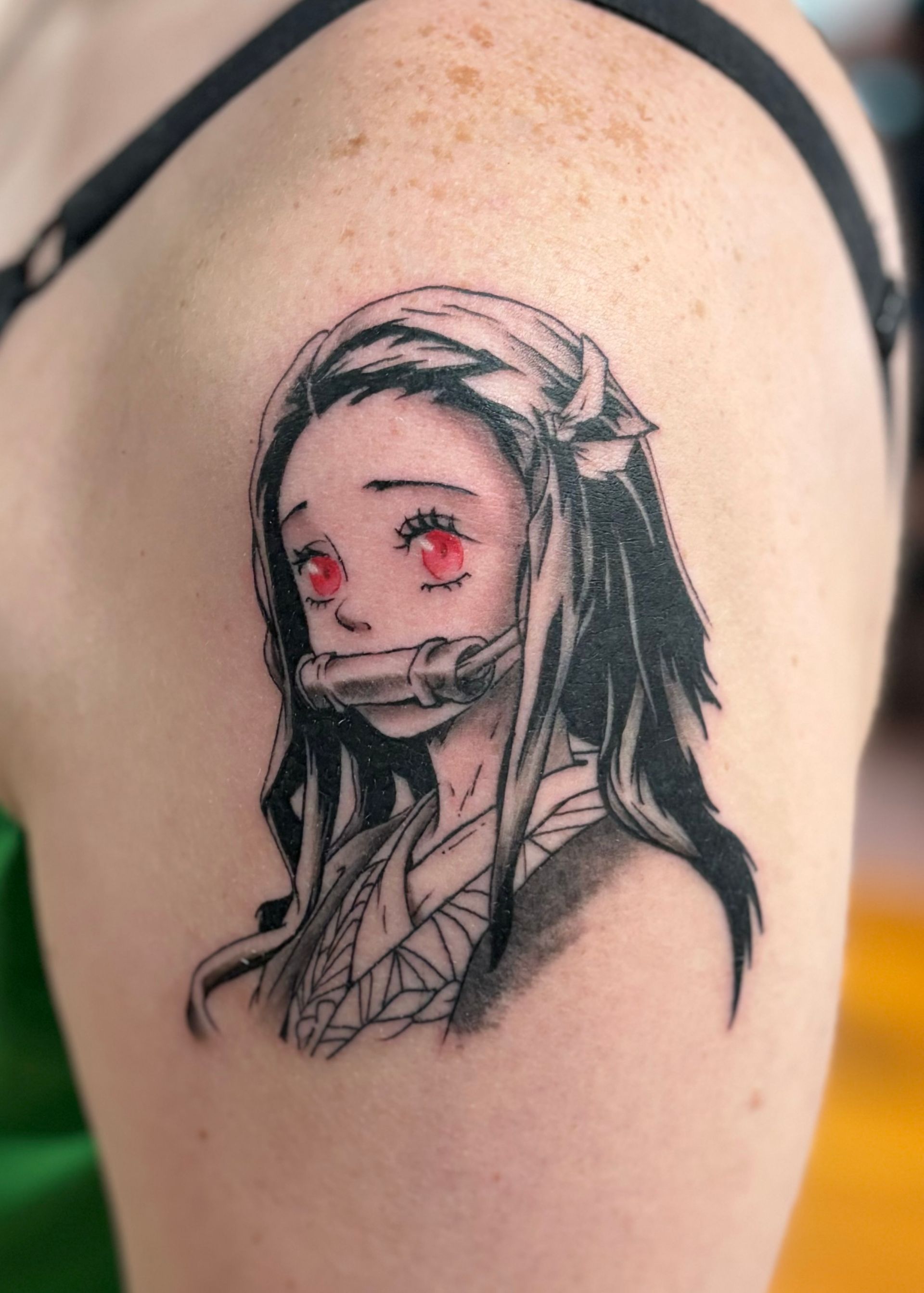 Tattoo of Nezuko from Demon Slayer. Black ink, red eyes, bamboo muzzle. On a person's shoulder.