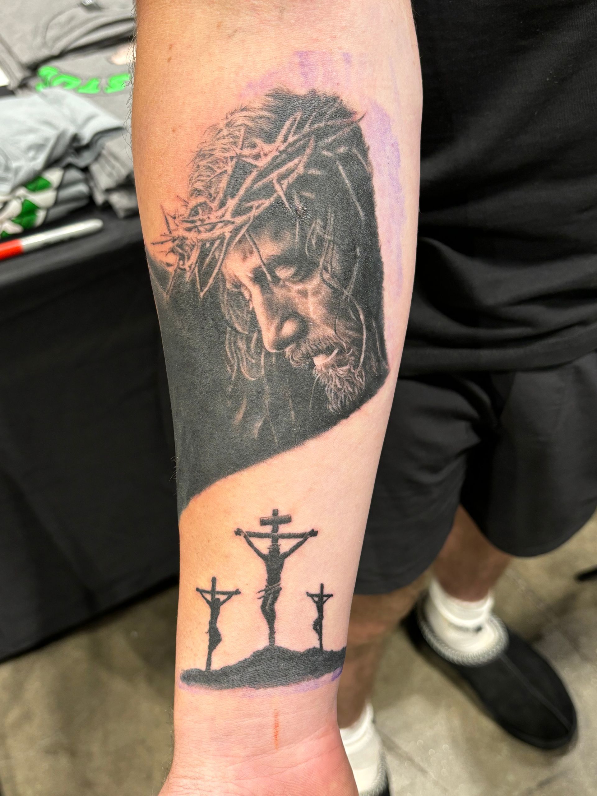 Tattoo of Jesus wearing a crown of thorns, with three crosses below on a wrist. Black and white.