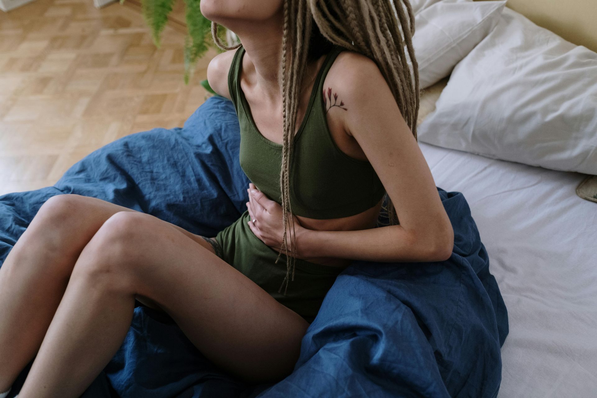 Woman with dreadlocks in green outfit, sitting on bed, holding stomach.