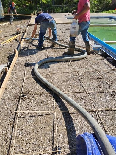 Workers pour concrete from a hose into a rebar-reinforced frame beside an outdoor swimming pool.