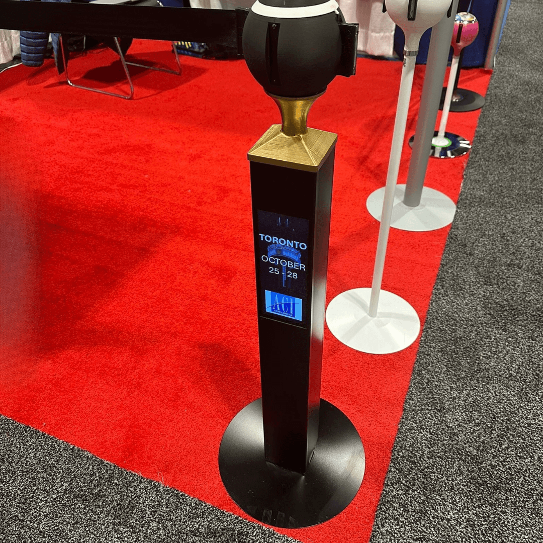 Een zwarte Square 100 afzetpaal met een klein videoschermpje toont een afbeelding. Op de achtergrond, op een rode loper, staan ​​nog andere afzetpalen.