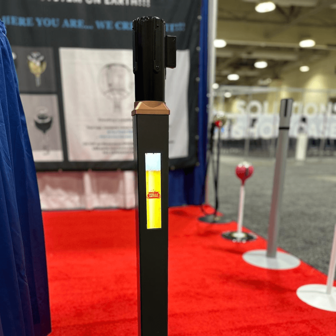 Zwarte Square 70 afzetpaal met gouden bovenkant en sticker, op een rode loper. Display op de achtergrond.