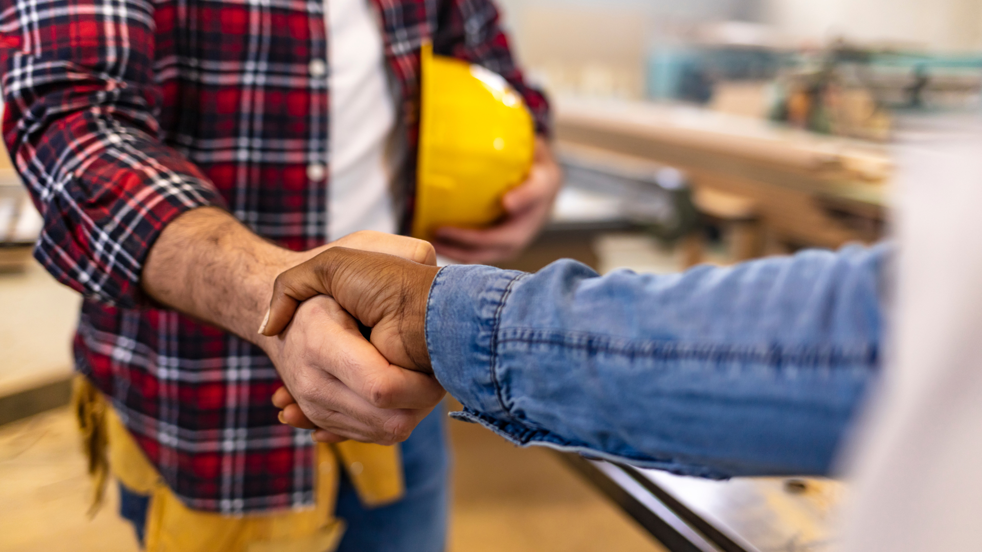 Two people shaking hands in a workshop; one in plaid shirt and hard hat, the other in denim.