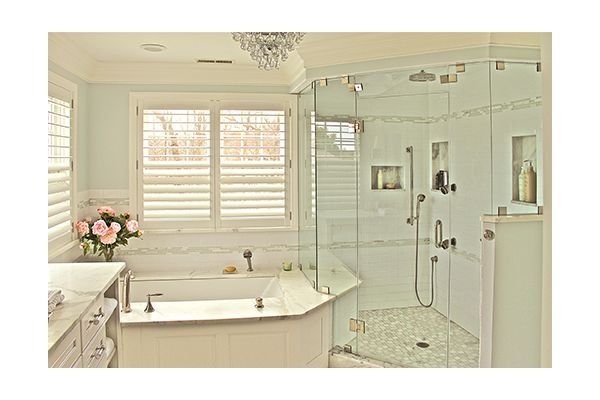 Elegant bathroom with white tile, glass shower, and soaking tub.  Window shutters and a chandelier add light.