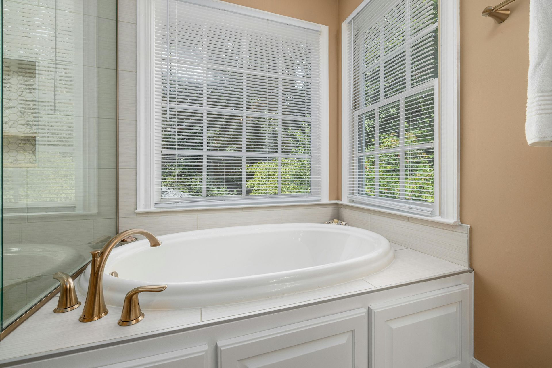 Oval bathtub in a bathroom, next to two windows with blinds. Gold faucet, white cabinetry and wall, beige walls.