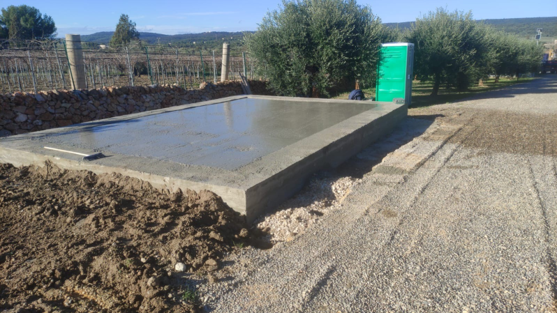 Bacino d'acqua in cemento a lato di una strada sterrata, con cumulo di terra e cabina di servizio verde in una zona rurale.