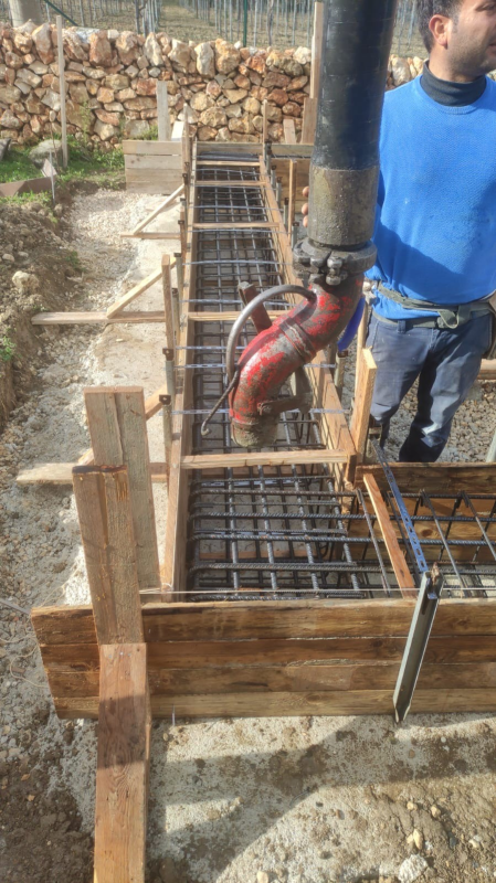 Operaio accanto a una trincea per casseforme in ferro in un cantiere edile, mentre pompa calcestruzzo con un tubo rosso.