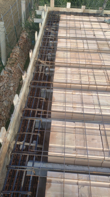 Armature in ferro e casseforme in legno posate per una soletta in cemento armato accanto a un muro e una recinzione in pietra.