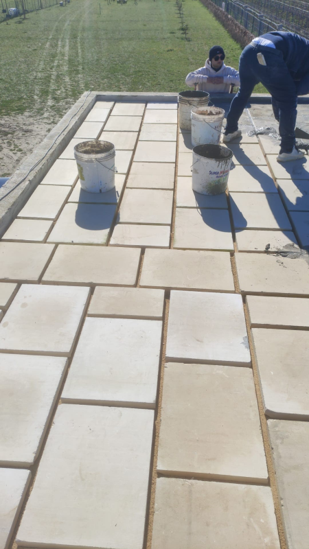 Operai che posano grandi lastre beige su un vialetto accanto a un campo erboso.