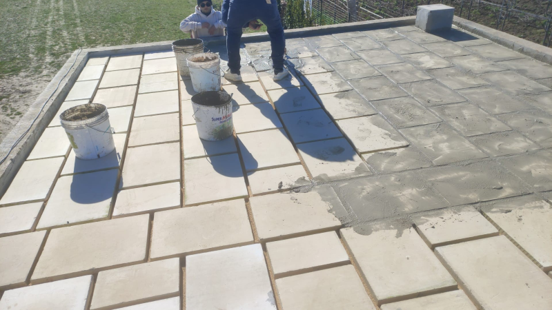 Area di lavoro sul tetto con secchi, una persona inginocchiata vicino a una porzione di cemento fresco e ombre proiettate dalla luce solare intensa.