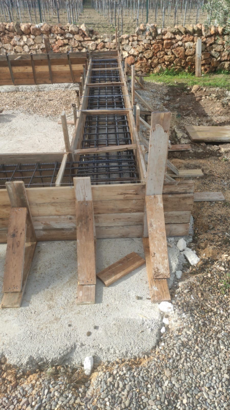 Cassaforma in legno attorno a una trincea per barre d'armatura in un cantiere edile, con un muro di ghiaia e pietra sullo sfondo.
